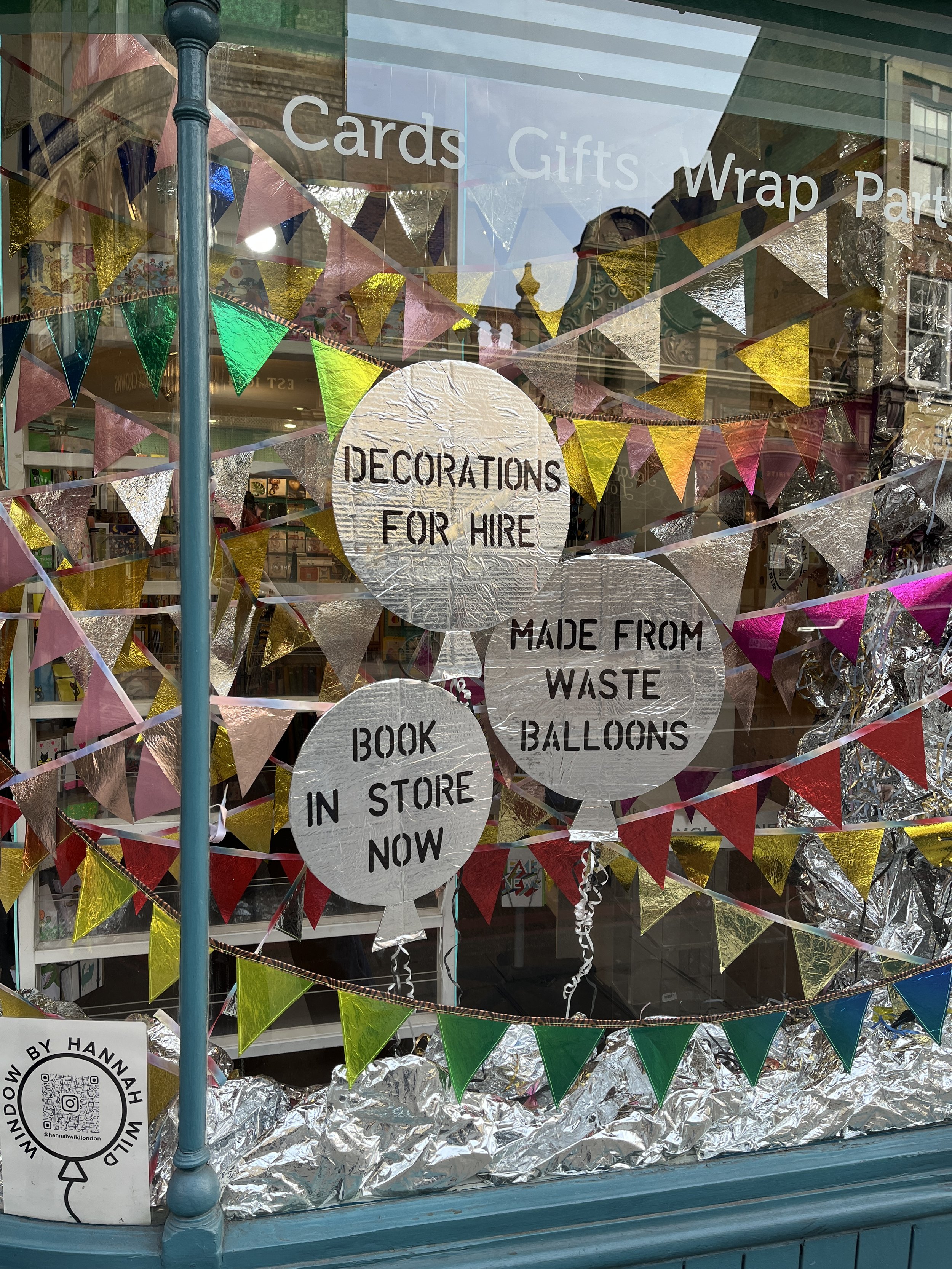 Store window decorated with colourful triangular banners and silver foil made from recycled foil balloons. Sign reads 'Decorations for Hire', 'Made from Waste', and 'Book in Store Now'.