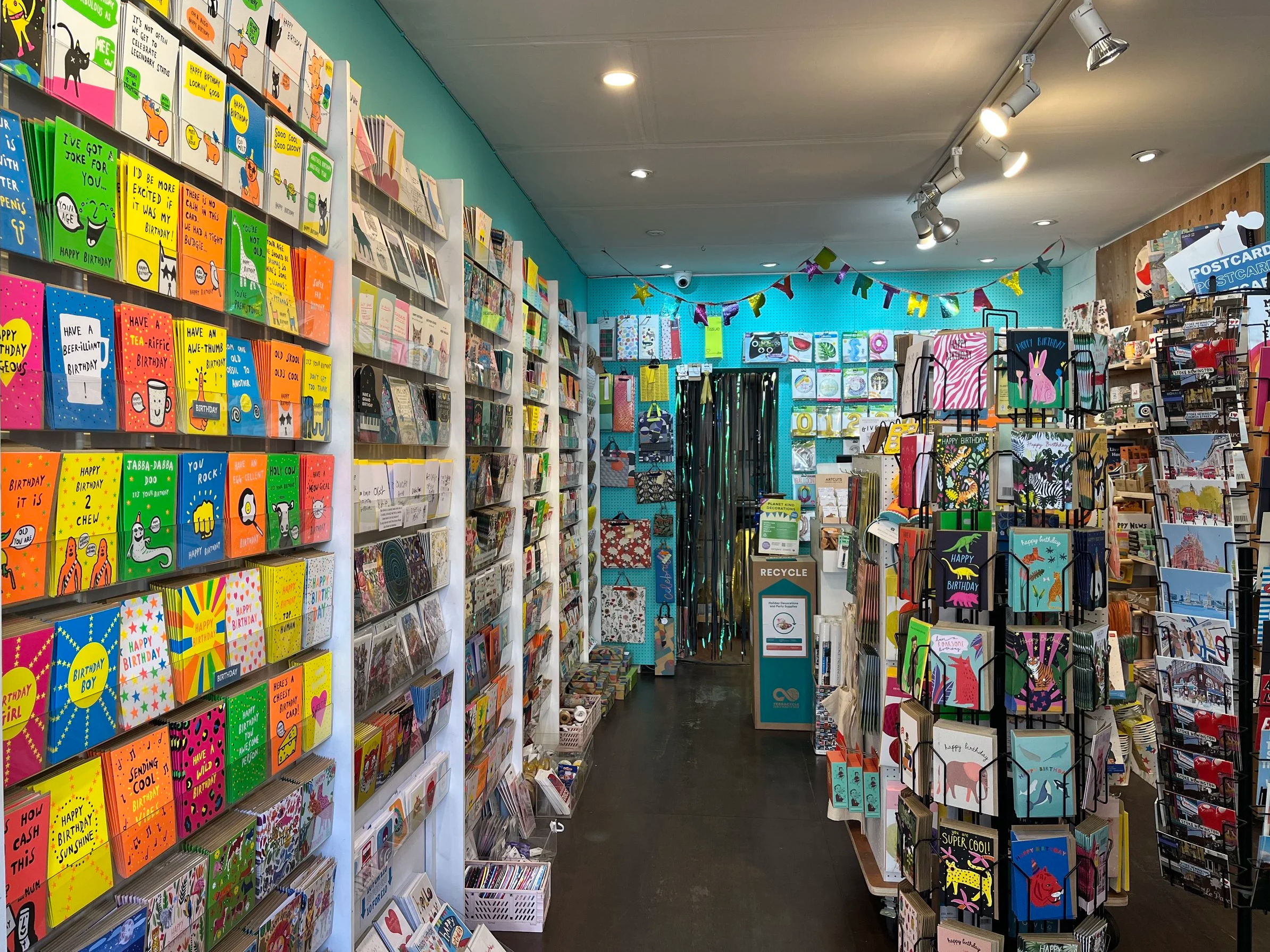 Inside a card store with colourful birthday cards on white shelves on the left and on display racks to the right, decorated with birthday banners hanging from the ceiling.
