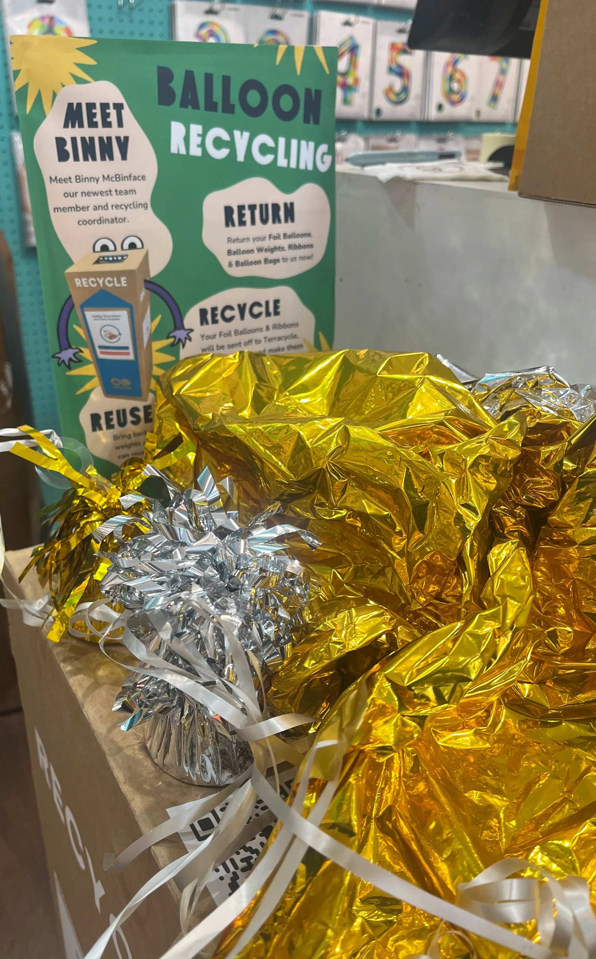 A display of crumpled metallic party balloons in gold and silver, placed in a cardboard box. In the background, a colorful informational sign about balloon recycling is visible.