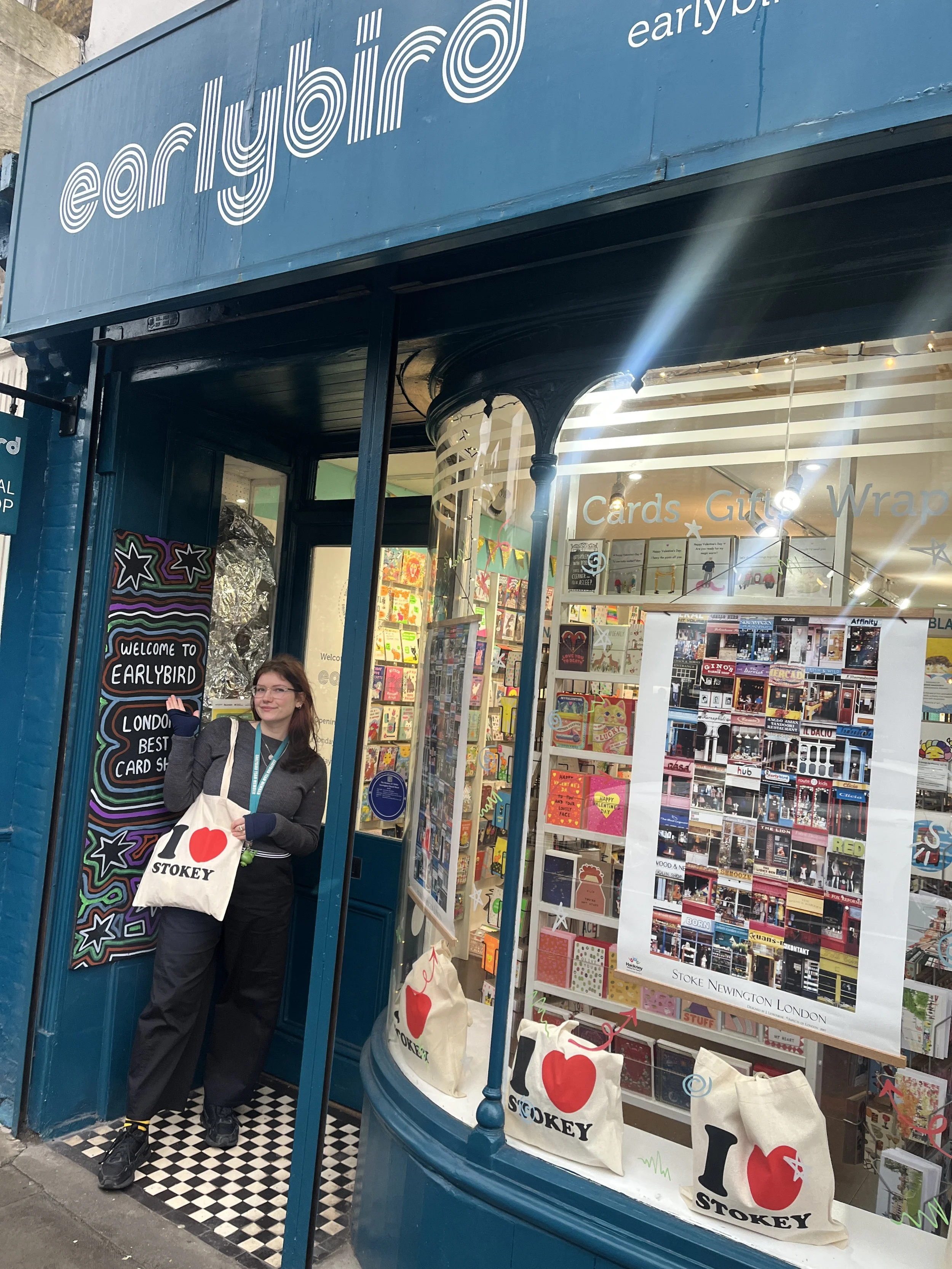 A woman standing outside a card and gift shop named 'earlybird'. She is holding a tote bag that says 'I ❤️ STOKEY' and is smiling at the camera. The shop window displays greeting cards and gifts.