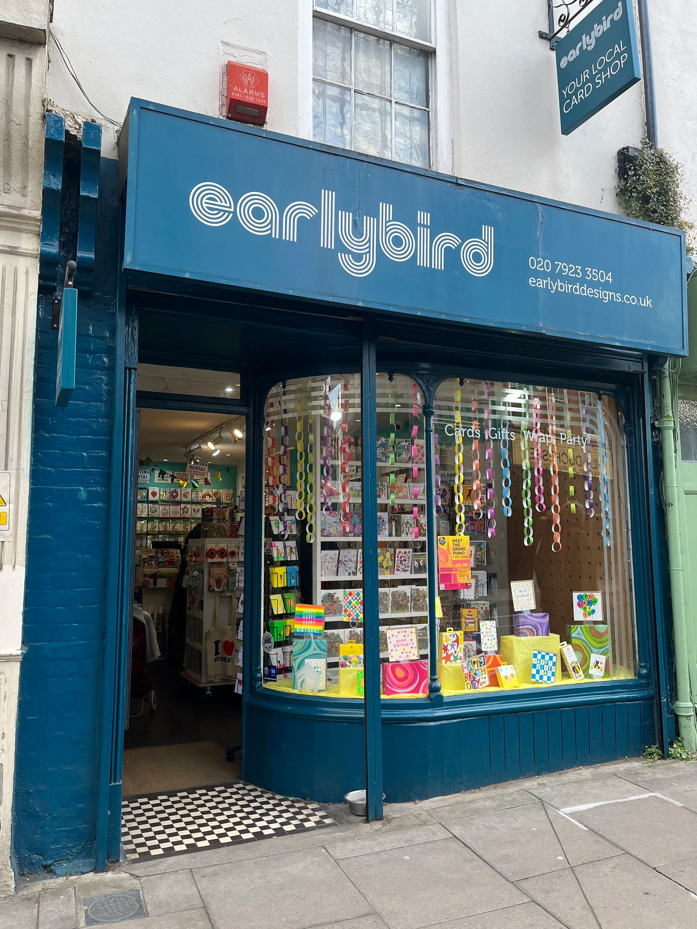 Storefront of a card shop called Earlybird Cards & Gifts, with a blue sign displaying the shop name and contact information. The window display features colourful greeting cards and gift wrap.