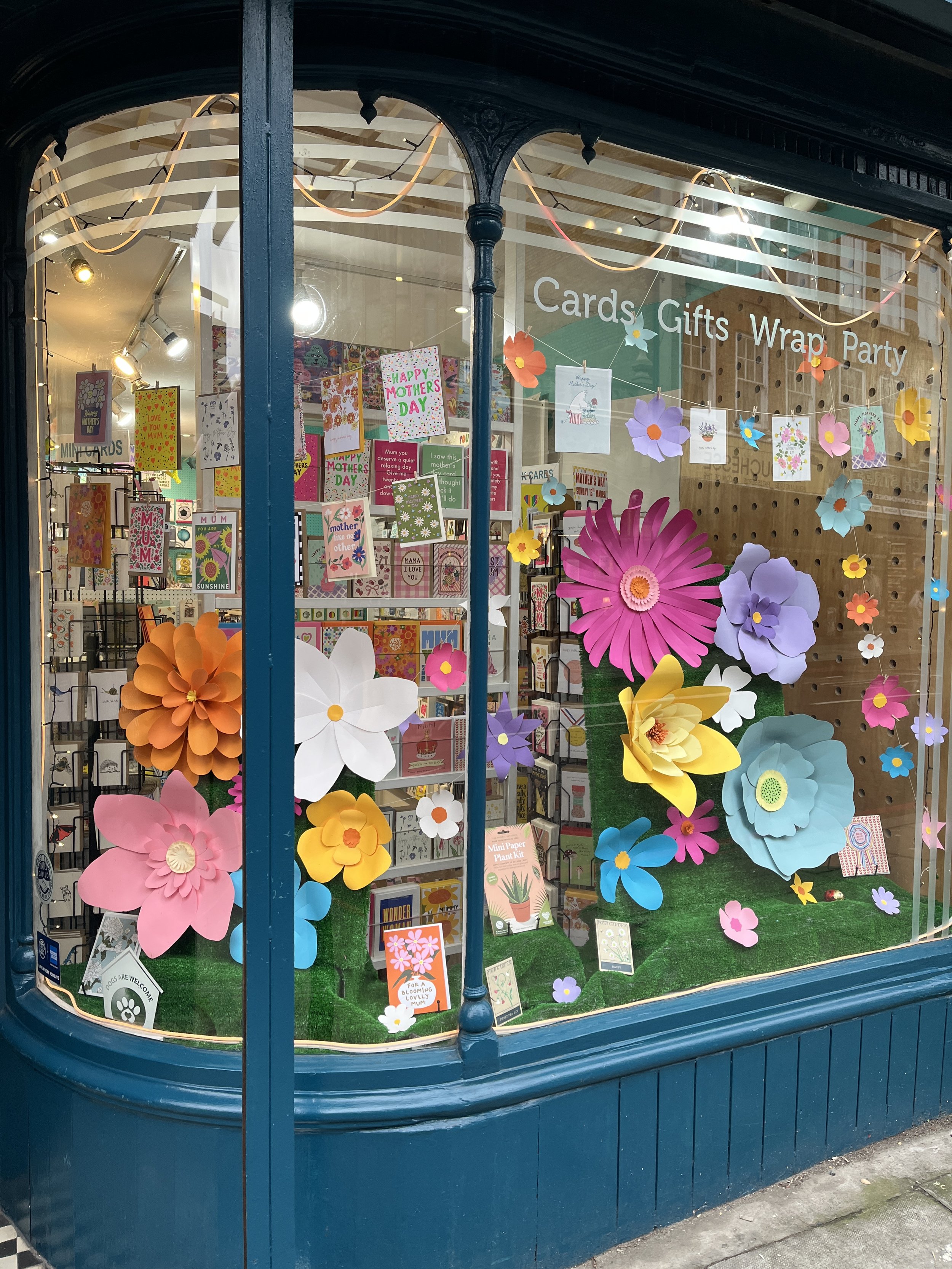 Store window showcasing Mother's Day greeting cards and colourful paper flowers decoration. The sign on the window reads 'Cards Gifts Wrap Party'.