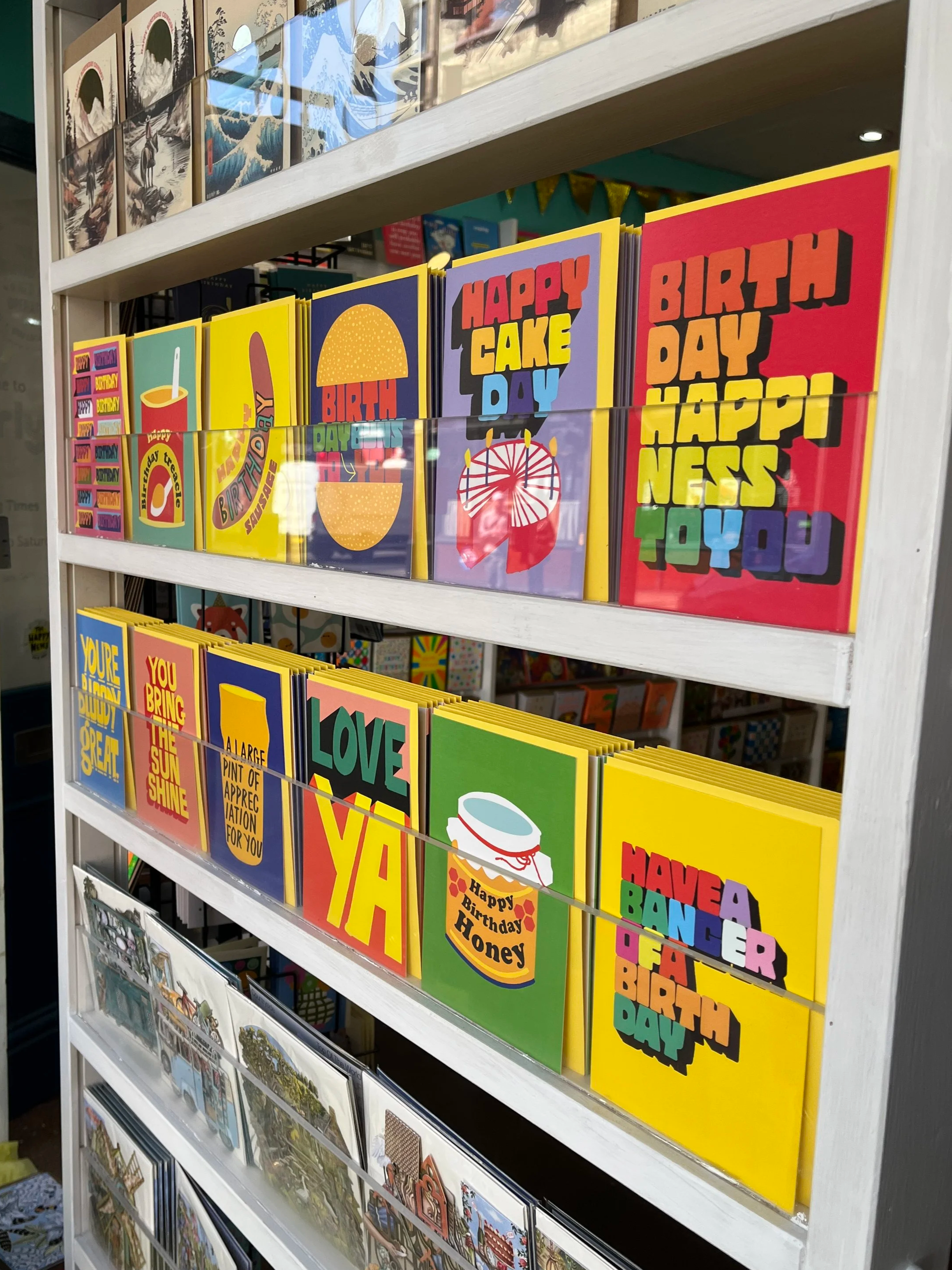 Store display shelf with colourful birthday greeting cards, featuring various designs and messages like "Happy Cake Day" and "Happy Birthday Honey".