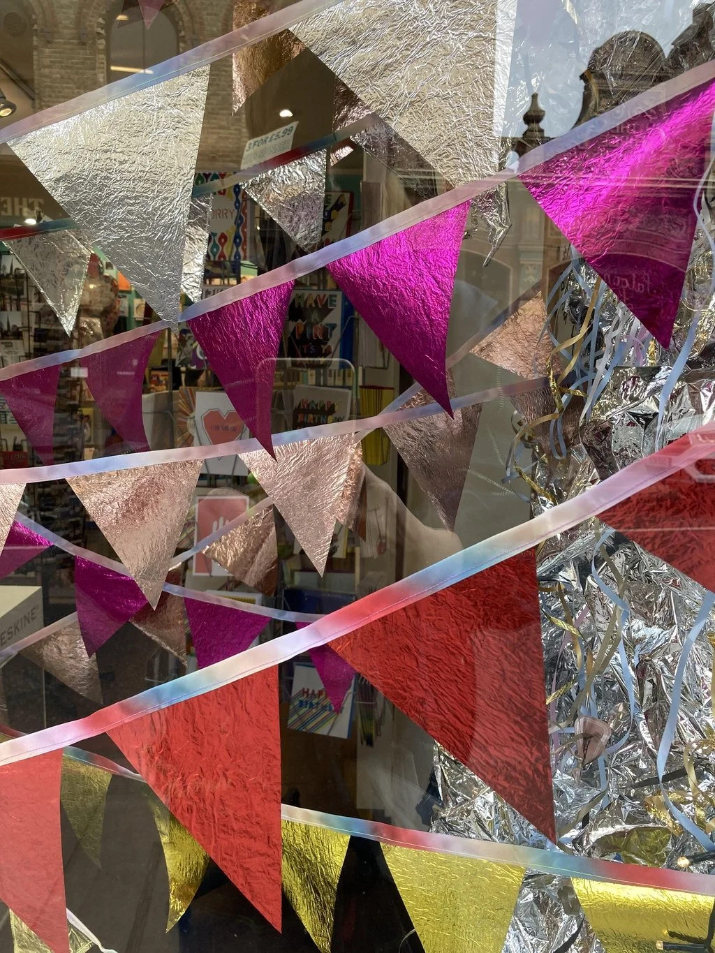 Colourful decorative bunting with pink, gold, silver, and red triangular flags hanging in front of a store window.