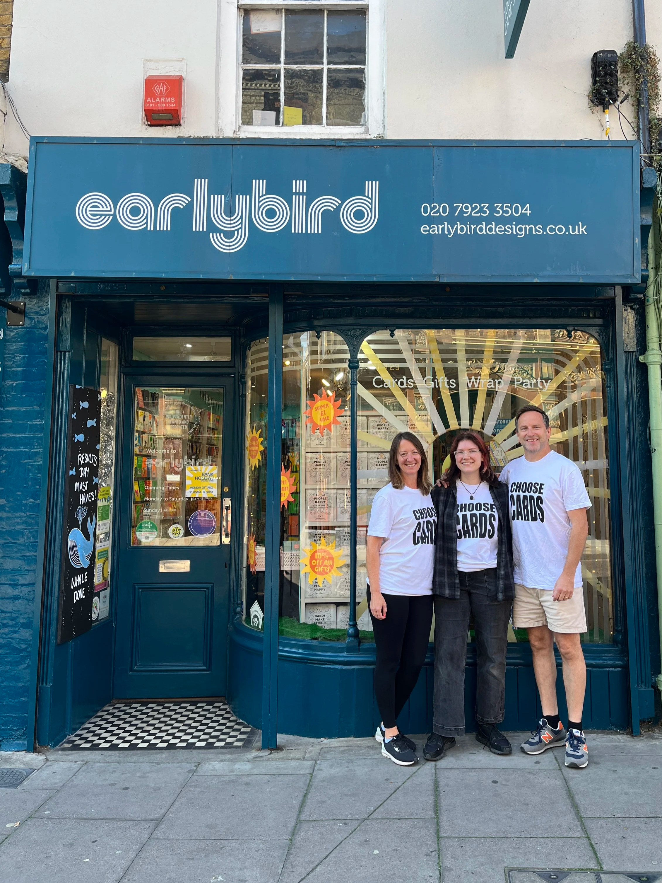 Three people standing outside a blue storefront with a sign for Earlybird, a card and gift shop, smiling for the camera.