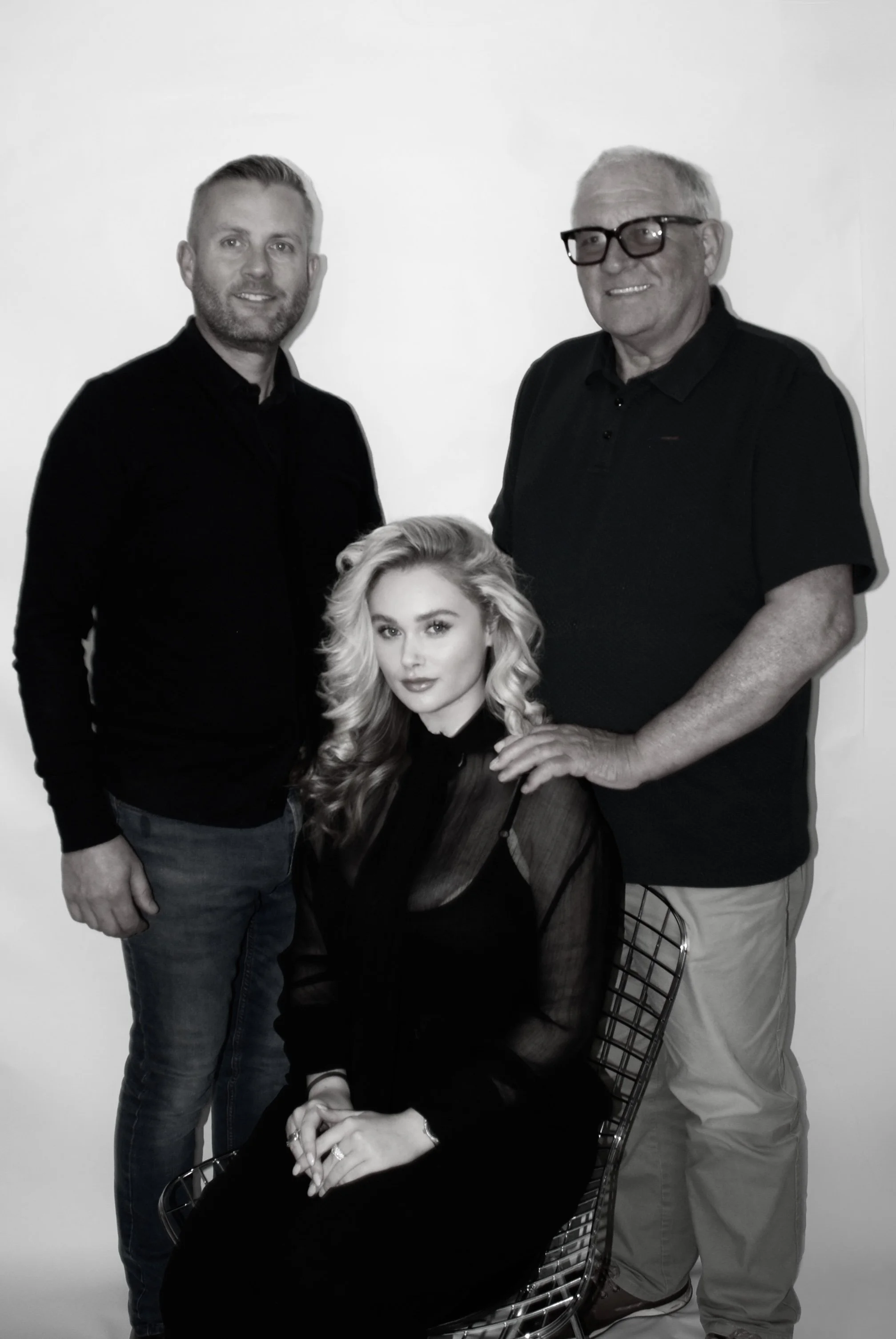 Four people, three men and one woman, posing together for a photo against a plain background. The woman is seated in a wire chair, with long wavy hair, wearing dark clothing, and looking directly at the camera. One man standing to her right has a beard, short hair, wearing a dark shirt and jeans. The other man standing to her left has gray hair, glasses, and is wearing a dark polo shirt and light-colored pants.