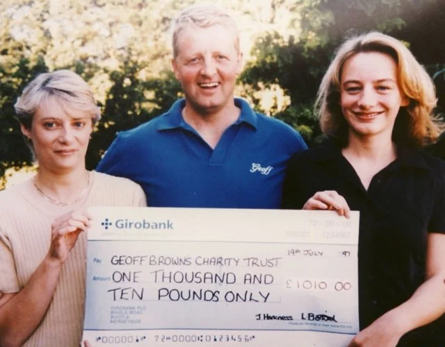 Three people standing outdoors in front of trees, holding a large check payable to Geoff Brown's Charity Trust for one thousand and ten pounds.