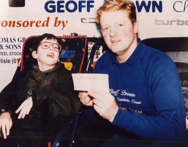 A young person in a powered wheelchair with glasses, wearing a black jacket, sitting next to a man with red hair in a blue shirt holding a check. They are at an event with a banner in the background.