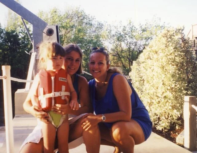 Three people, an adult woman and two children, sitting on a deck with trees in the background. The child on the left is wearing a life jacket and swimsuit. The woman and the other girl are smiling at the camera.