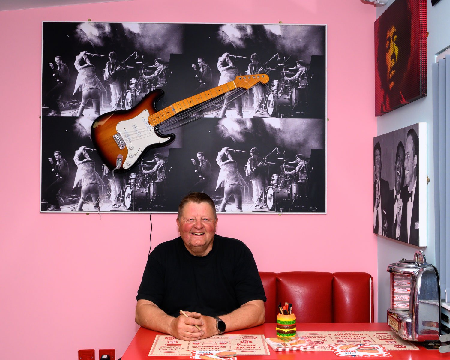 Geoff Brown smiling  sitting at a red table in a pink-walled diner. Behind him is a black and white picture of a band playing on stage, with a guitar mounted on the wall.