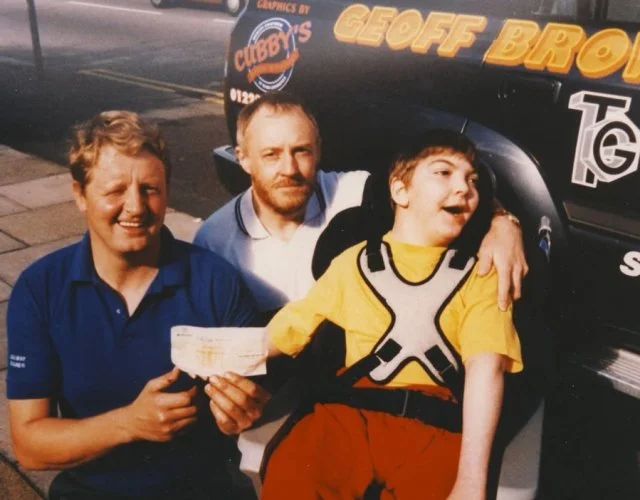 Three men posing outdoors near a van. The man on the left is holding a check or piece of paper. The van displays the text 'Cubby’s Graphics by Geoff Brown'.