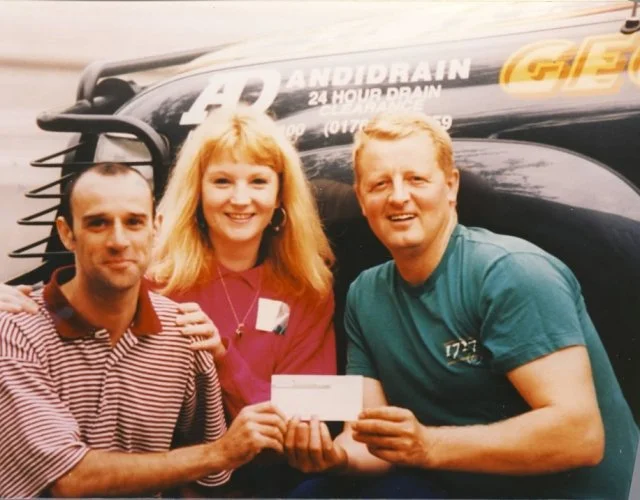 Three people, two men and one woman, smiling and holding a check in front of a black truck with advertising. The woman has long blonde hair and is wearing a pink top. The man on the left has short dark hair and is wearing a red and white striped shir