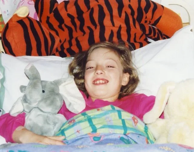 Child lying in bed with stuffed animals, including a gray rabbit, a yellow duck, and a tiger plush, smiling.