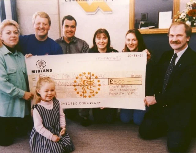 Group of seven people, including a young girl in the foreground, holding a large check for 500 pounds made out to Geoff Brown, in an office or community center setting.