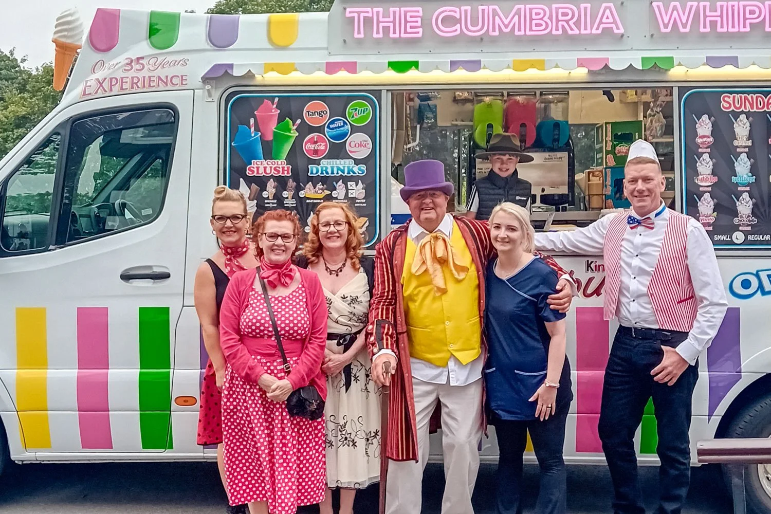 A group of six people in colorful costumes posing in front of a vibrant ice cream truck, with two children working inside the truck. The truck has ice cream and drink advertisements and rainbow-colored stripes.
