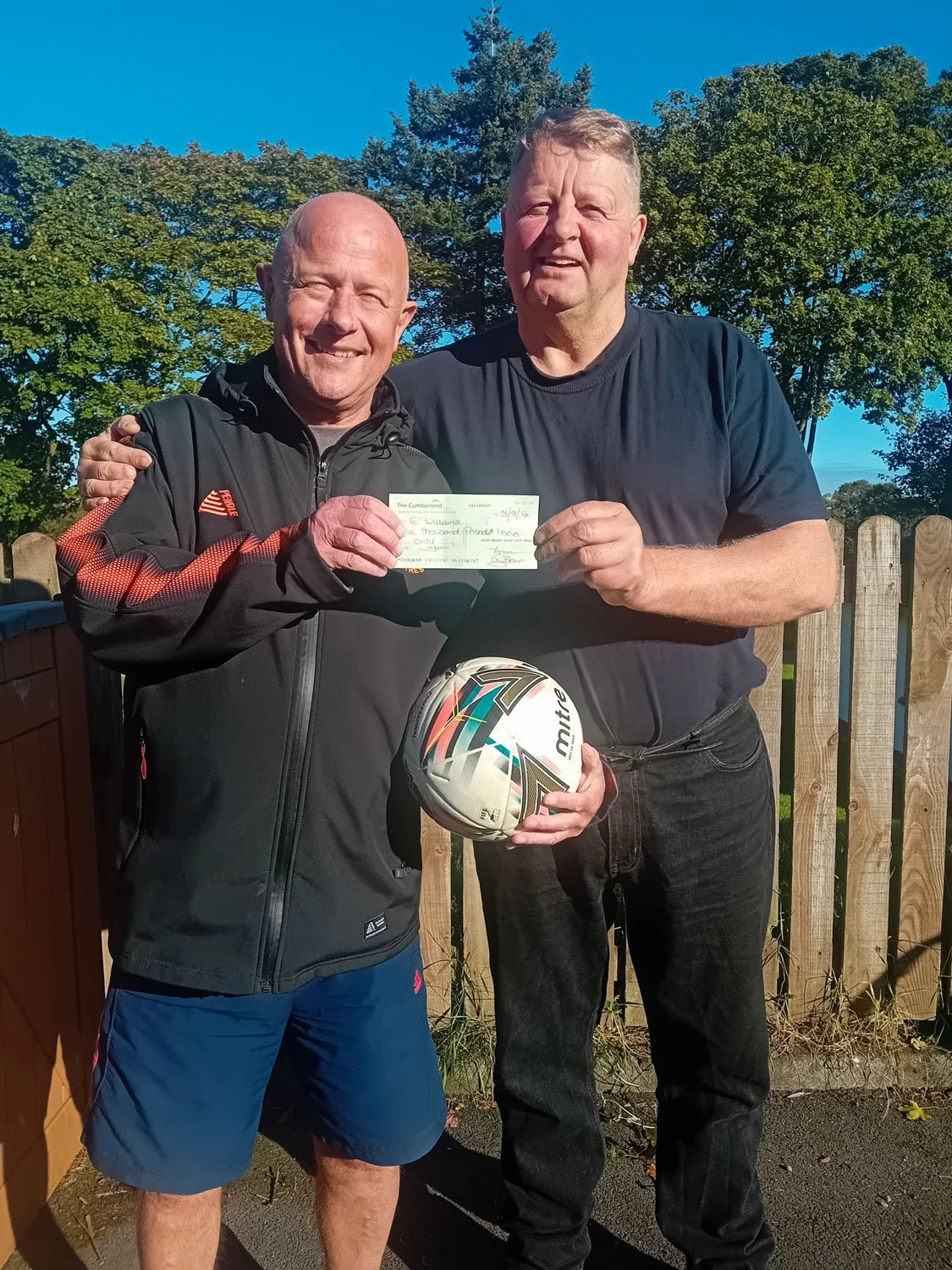 Two men outdoors holding a check and a soccer ball, smiling, with trees and a wooden fence in the background.