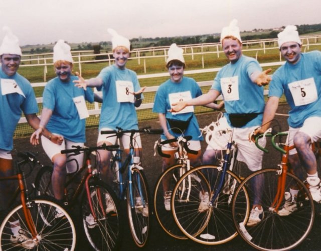 A group of six people dressed as cupcakes, wearing blue shirts, white hats, and numbered bibs, standing with bicycles on a race track.
