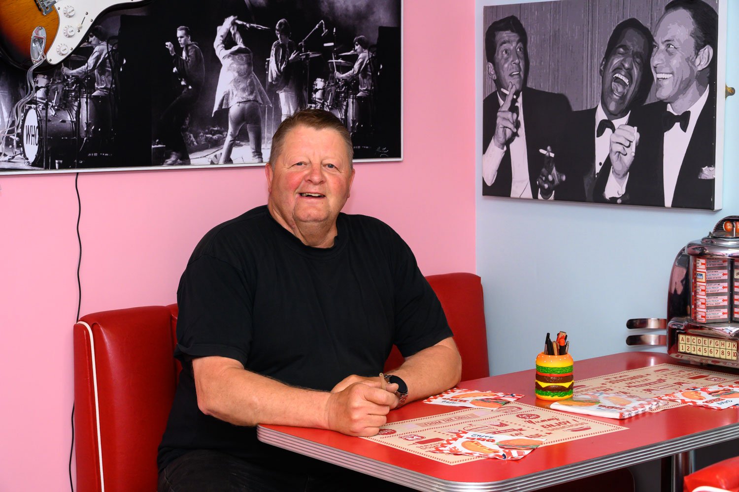 Geoff Brown sitting at a red booth in a retro diner, smiling and wearing a black shirt. The decor includes black and white photos of musicians on the wall behind him and a colorful burger-shaped pencil holder on the table.