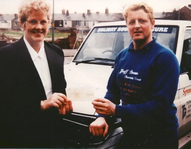 A woman with glasses and a suit handing a cheque to a man in a blue sweatshirt standing in front of a white van with 'SOLWAY SQUARE' written on the windshield.