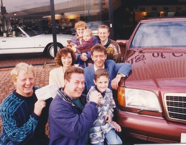 A group of seven people, including five adults and two children, standing outside a car dealership in front of a red SUV. They are smiling and appear to be celebrating a car purchase.