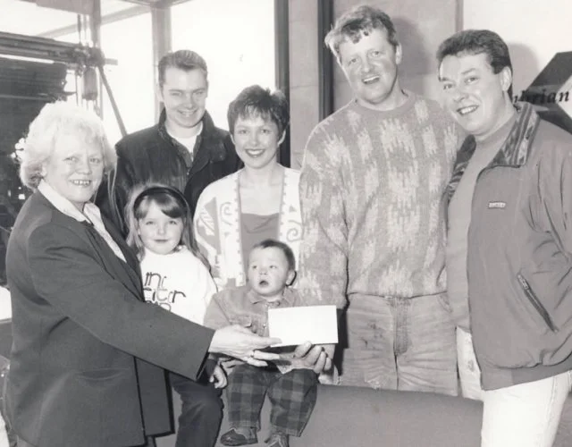 A group of six adults and two children standing together holding together the cheque from Geoff Brown Charity Trust