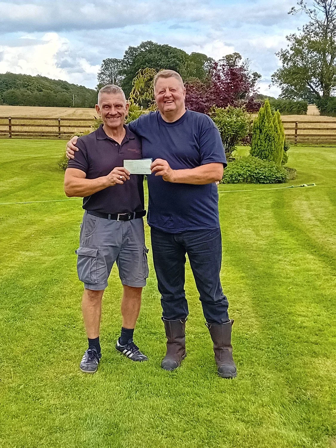 Two men standing on a lawn, smiling, one holding a check, with trees, bushes, and a wooden fence in the background.