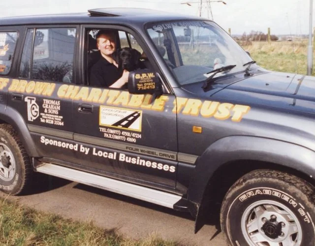 A man sitting in a black van with charity and local business sponsorship decals, smiling and waving at the camera, in an outdoor rural setting.