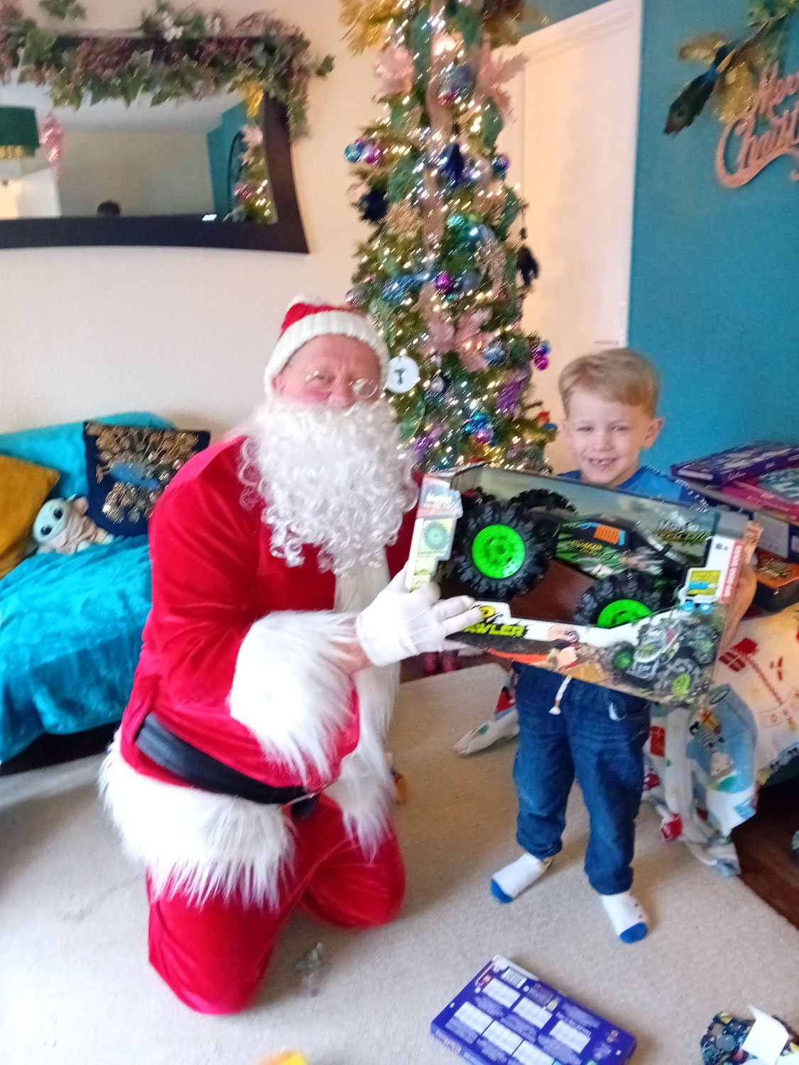 Santa Claus, dressed in a red suit with white fur trim, kneeling next to a young boy holding a toy truck, in a room decorated for Christmas with a decorated Christmas tree, wrapped presents, and holiday decorations.