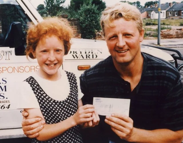 A young girl with curly red hair and a man with short blond hair are smiling and holding a check together outdoors, with a white van and trees in the background.