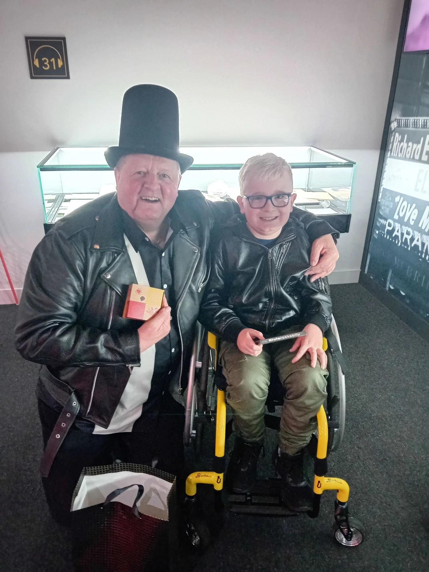 Two men, one older with a tall black hat and a leather jacket, and a young man with glasses and a leather jacket, sitting together with a wheelchair, smiling at the camera indoors.