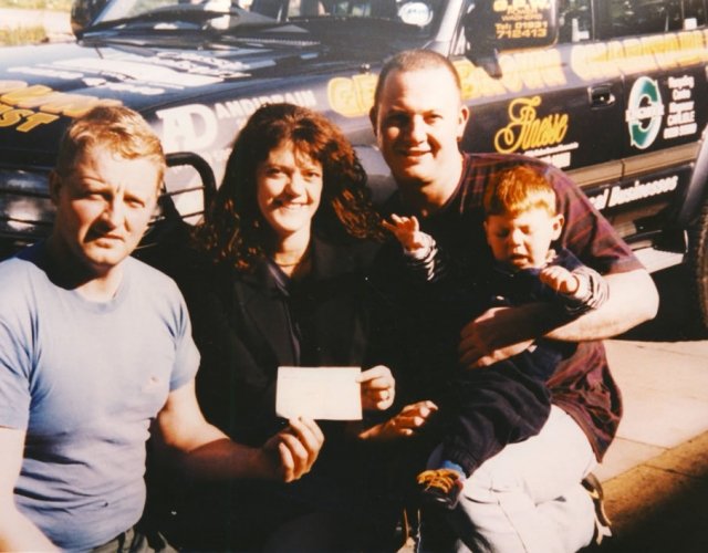 Four people standing outdoors in front of a race car, with a woman holding a small white object, possibly a check or a card, and a young boy sitting on a man's lap.
