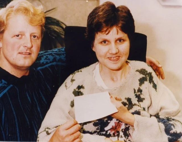 A woman sitting in a chair holding a piece of paper, with a man sitting next to her and resting his arm on her shoulder, both smiling at the camera.