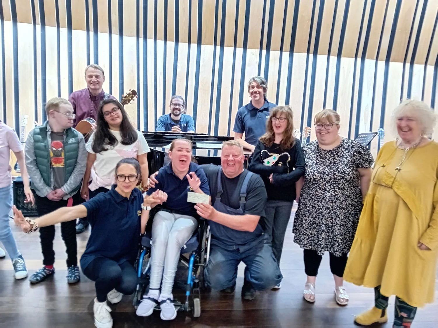 Geoff Brown and group of people, some smiling and laughing, in a music rehearsal or performance space, with some holding musical instruments and a piano visible in the background. Geoff giving a cheque from Geoff Brown Charitable Trust