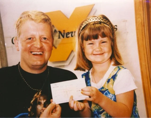 A man and a young girl smiling, holding a check in front of a news station sign.