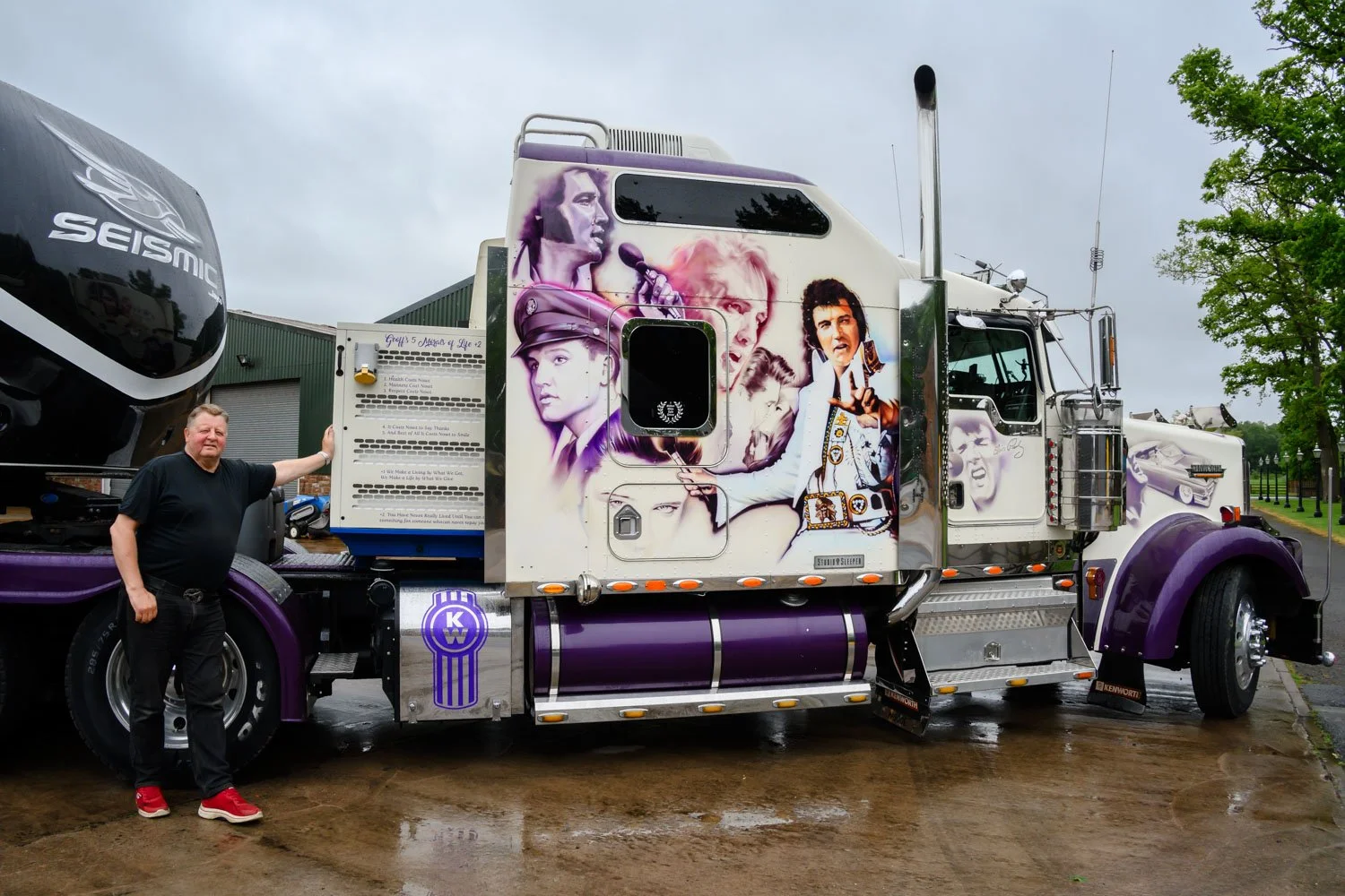 A man standing next to a large semi-truck with movie and music icons painted on its side, including images of Freddie Mercury, Lady Gaga, and other performers, in an outdoor setting with green trees and cloudy sky.