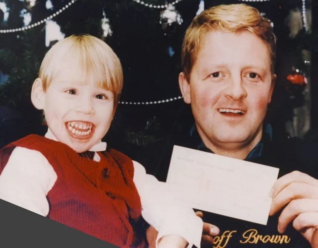 A young boy and an adult man smiling, holding a check, in front of a decorated Christmas tree.