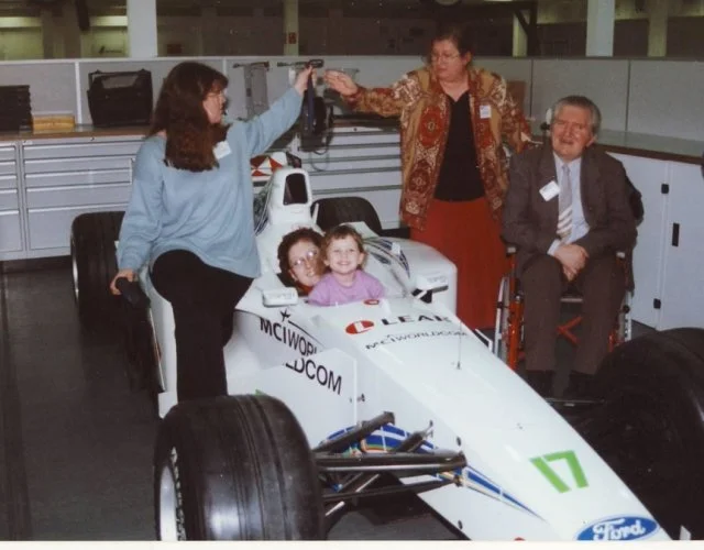 A young girl and a woman sitting in a white Formula racing car in an indoor garage, with two women and a man nearby. The woman standing on the left is reaching out and high-fiving another woman, who is standing in front of her. The man is sitting in 