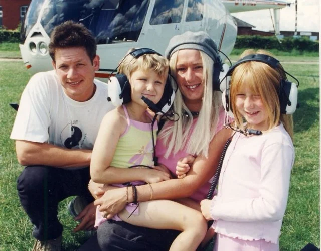A woman and three children wearing headsets with microphones, sitting on grass in front of a helicopter.