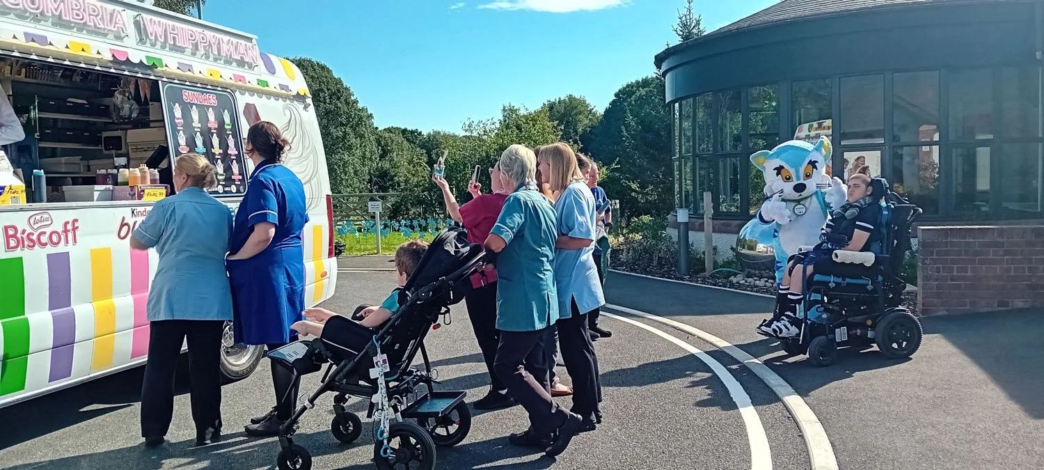 People in medical uniforms and caregivers at an ice cream truck, with a mascot character in front of a building.