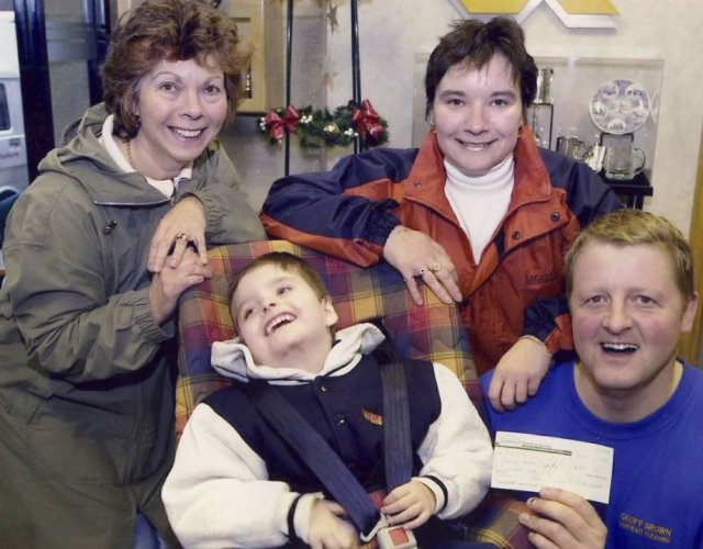 Four people smiling in a cozy indoor setting; a woman and a man standing behind two boys, one holding a check, with festive decorations in the background.