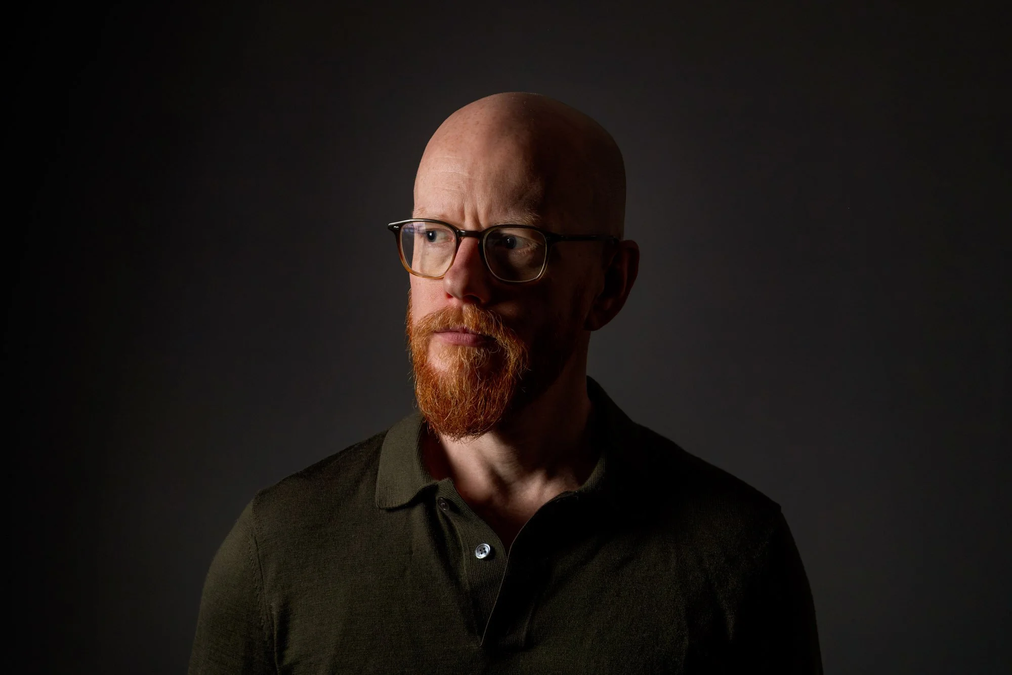A man with glasses and a red beard wearing a dark shirt looks to the side against a dark background.