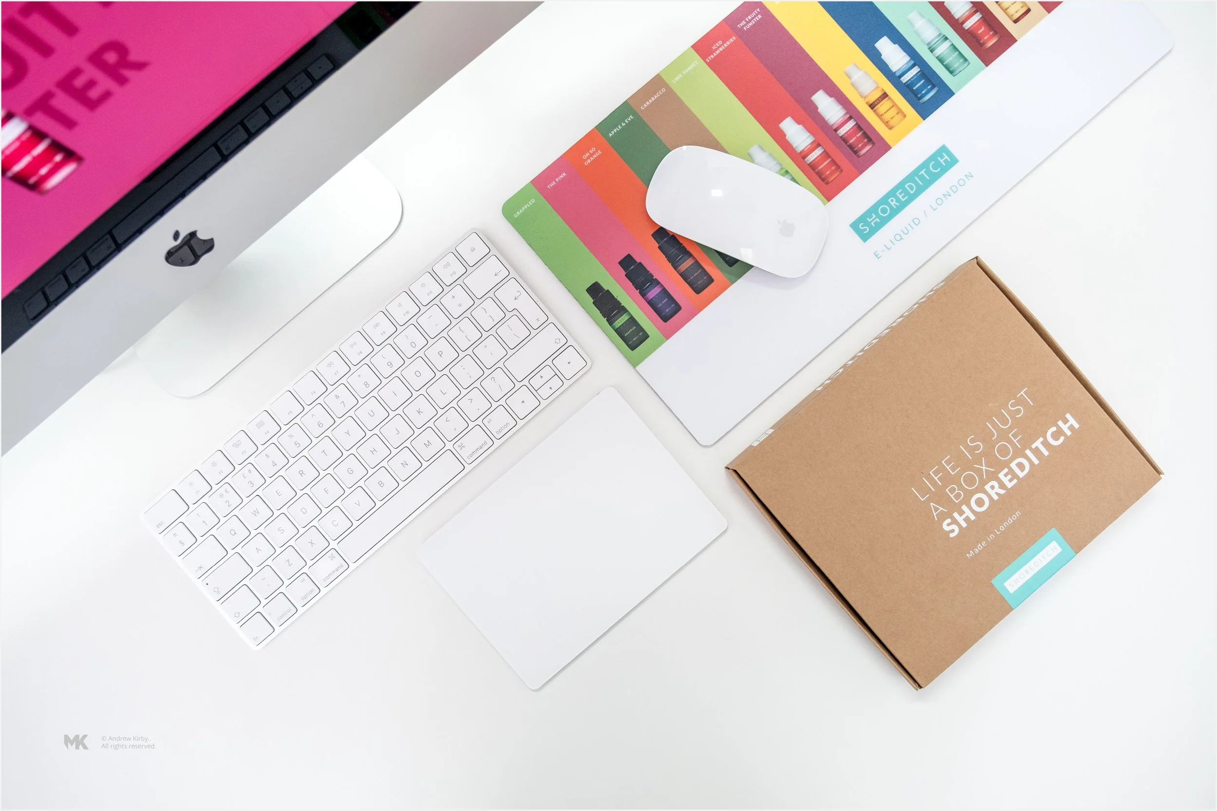 Overhead view of a white desk with an Apple iMac computer, a wireless keyboard, a wireless mouse, a colorful brochure for Shoreditch e-liquid, and a cardboard box labeled 'Life is just a box of Shoreditch'.