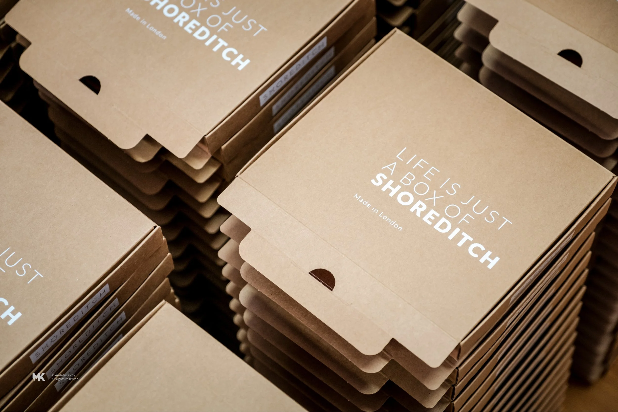 Stacks of cardboard boxes with white printed text that says, "Life is just a box of Shoreditch," indicating they are made in London.