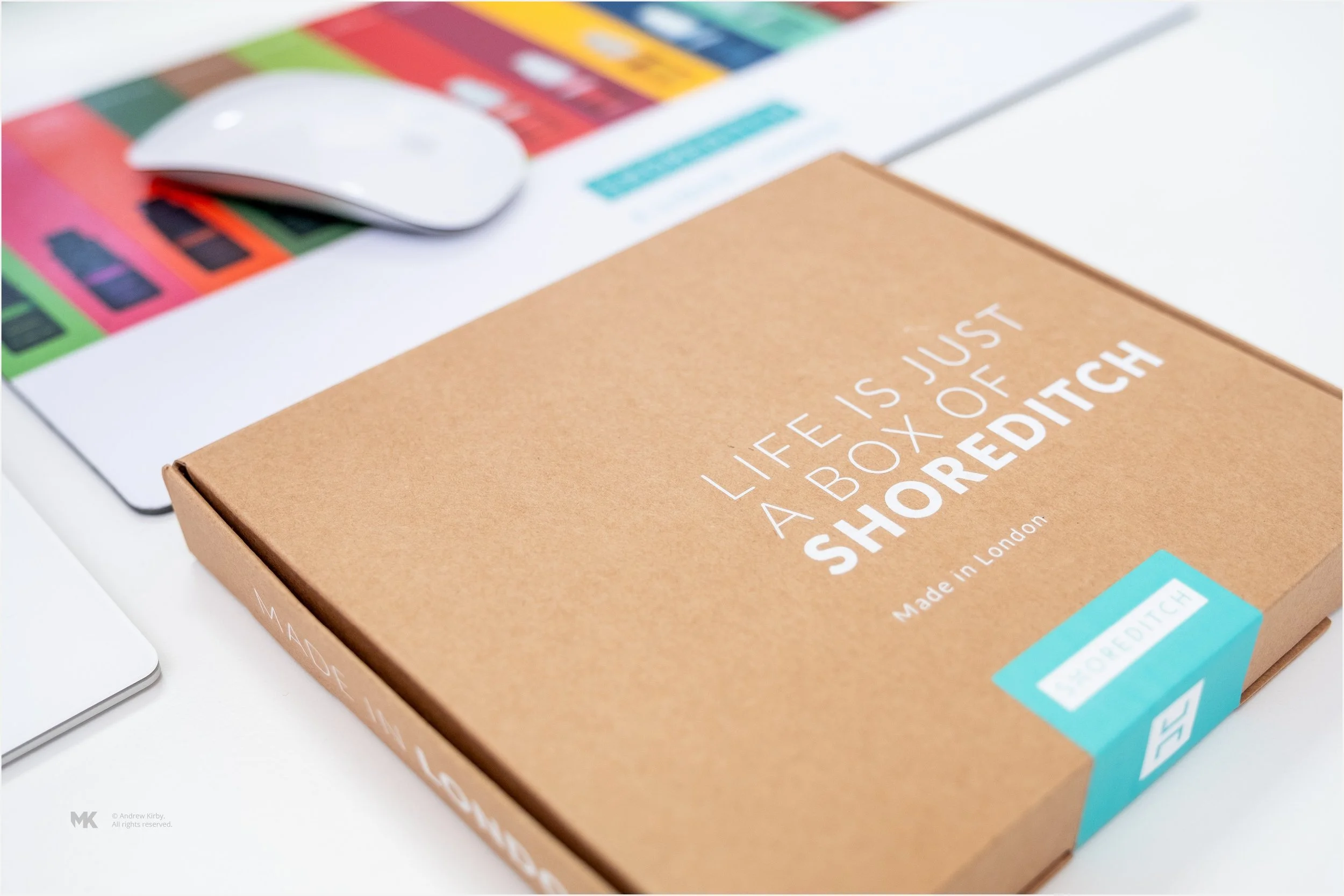 A brown cardboard box with the quote 'Life is just a box of shortedch' written on it, placed on a white desk with a computer mouse and a colorful chart or product packaging in the background.