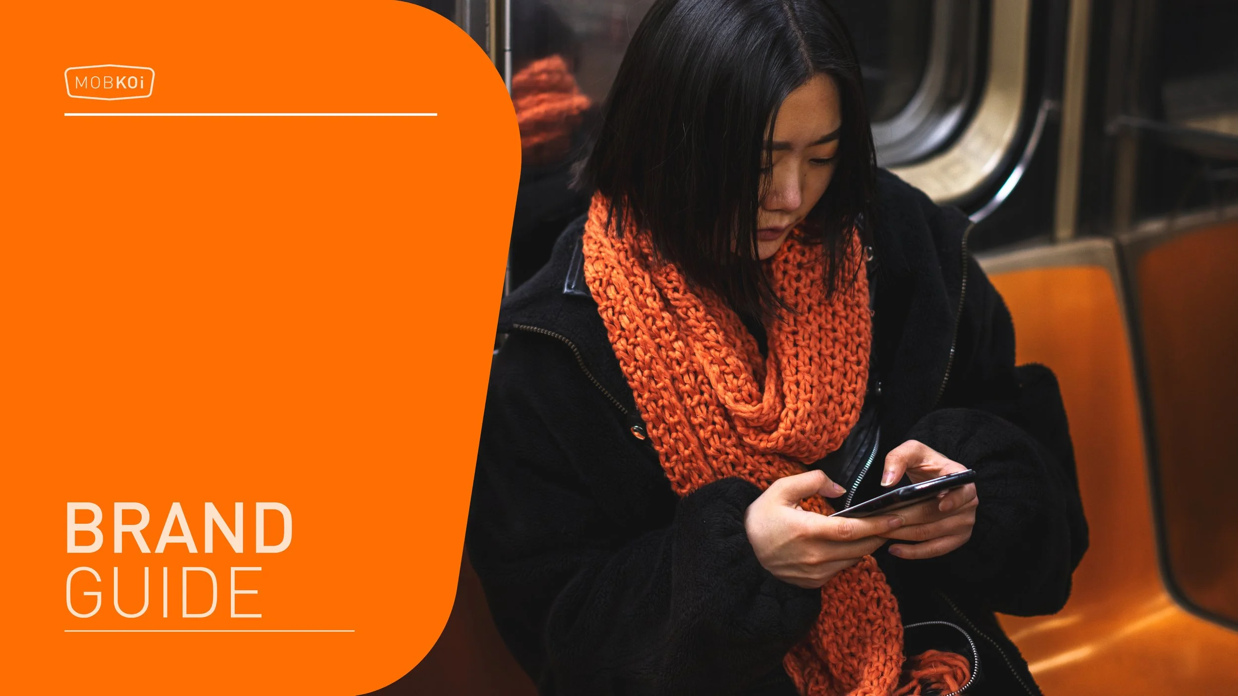 A woman in a black jacket and orange knitted scarf using her phone while sitting on an orange bench in a subway station.