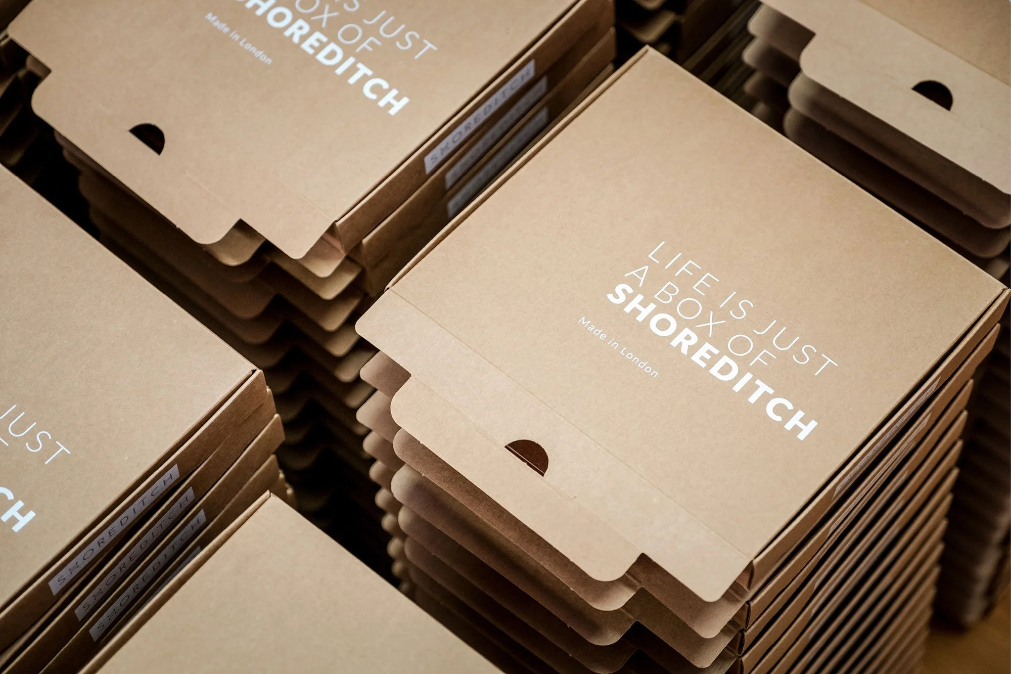 Stacks of brown paper boxes with white text stating "LIFE IS JUST A BOX OF SHORTEDITCH" and "Made in London," arranged on a wooden surface.