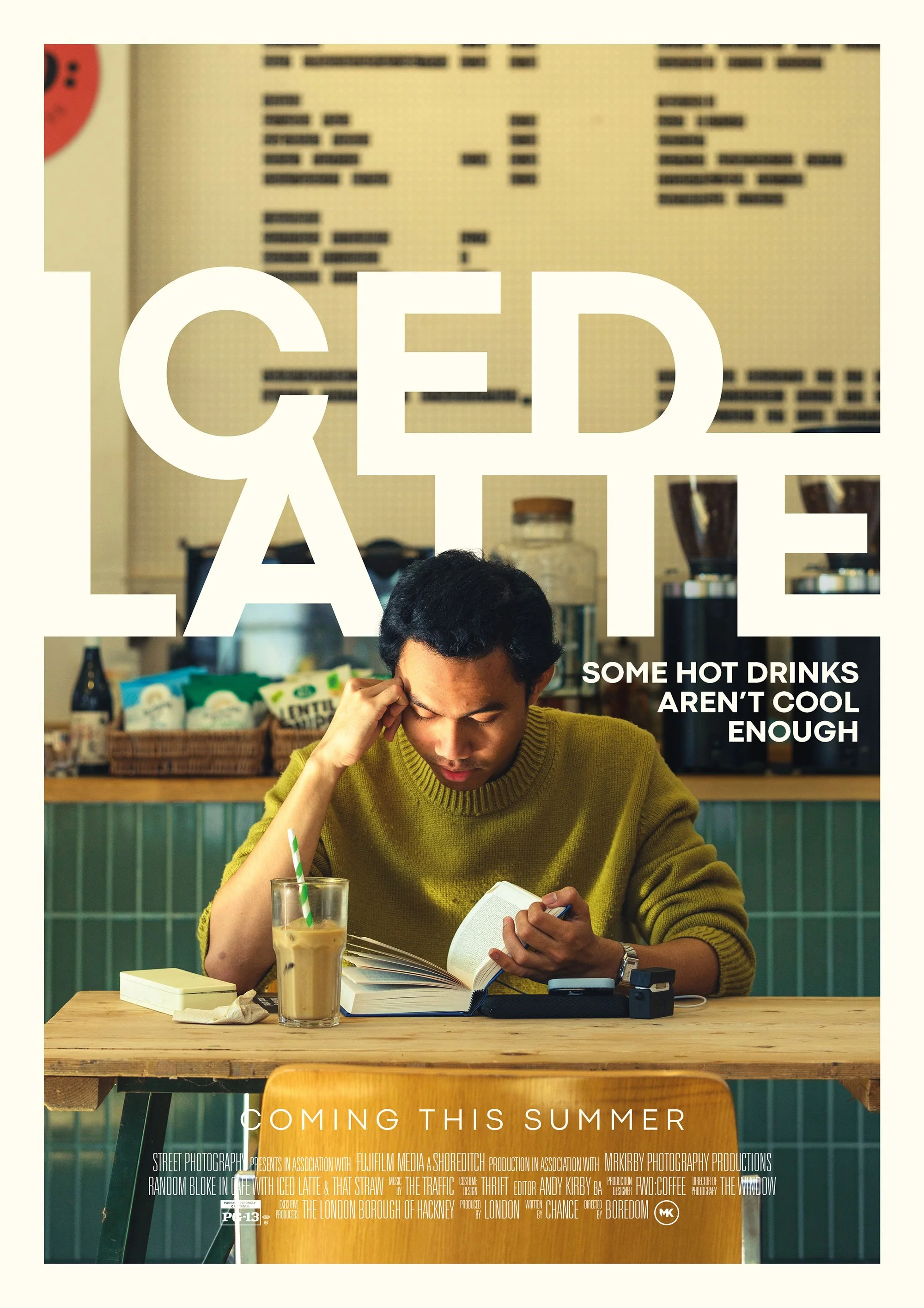 A man in a mustard yellow sweater sitting at a wooden table in a cafe, reading a book, with a tall glass of iced coffee in front of him. The background shows a menu on the wall and various snacks on shelves.