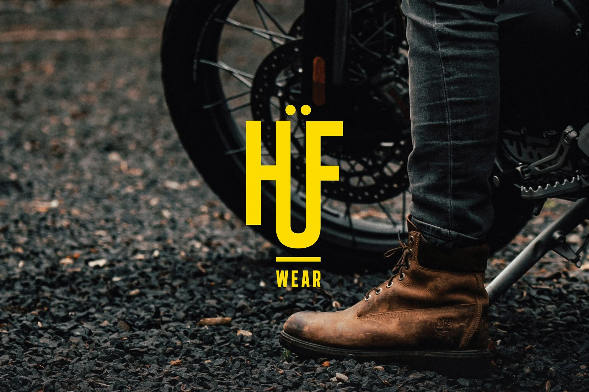 Close-up of a person's lower leg wearing brown leather boots and dark jeans, standing next to a bicycle on a gravel surface, with yellow text overlay that says 'HÖ WEAR'.