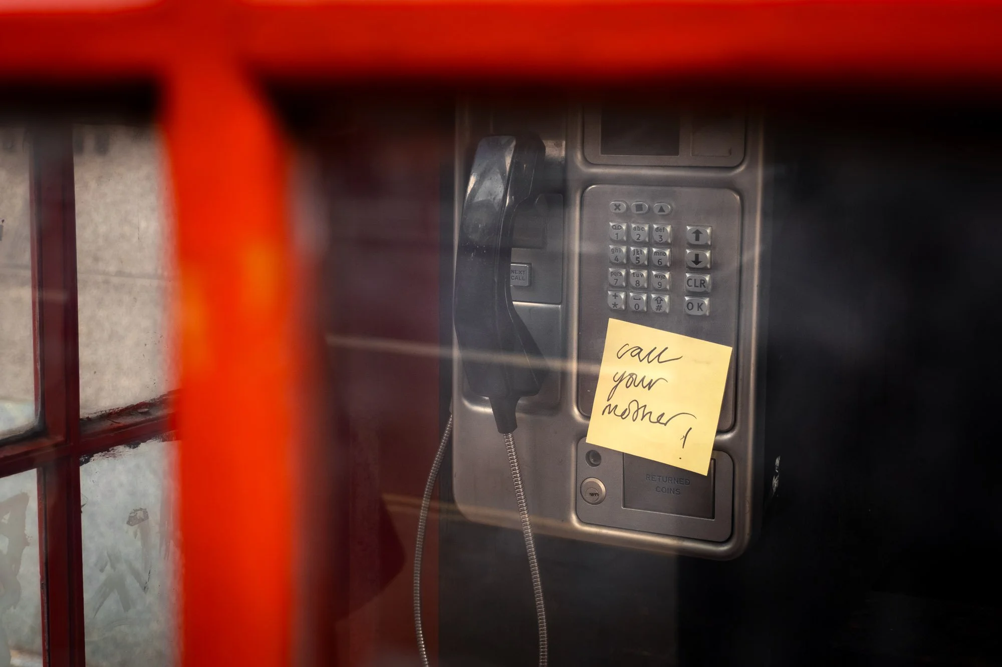 A public payphone on a wall with a yellow sticky note attached that says 'call your mom.'