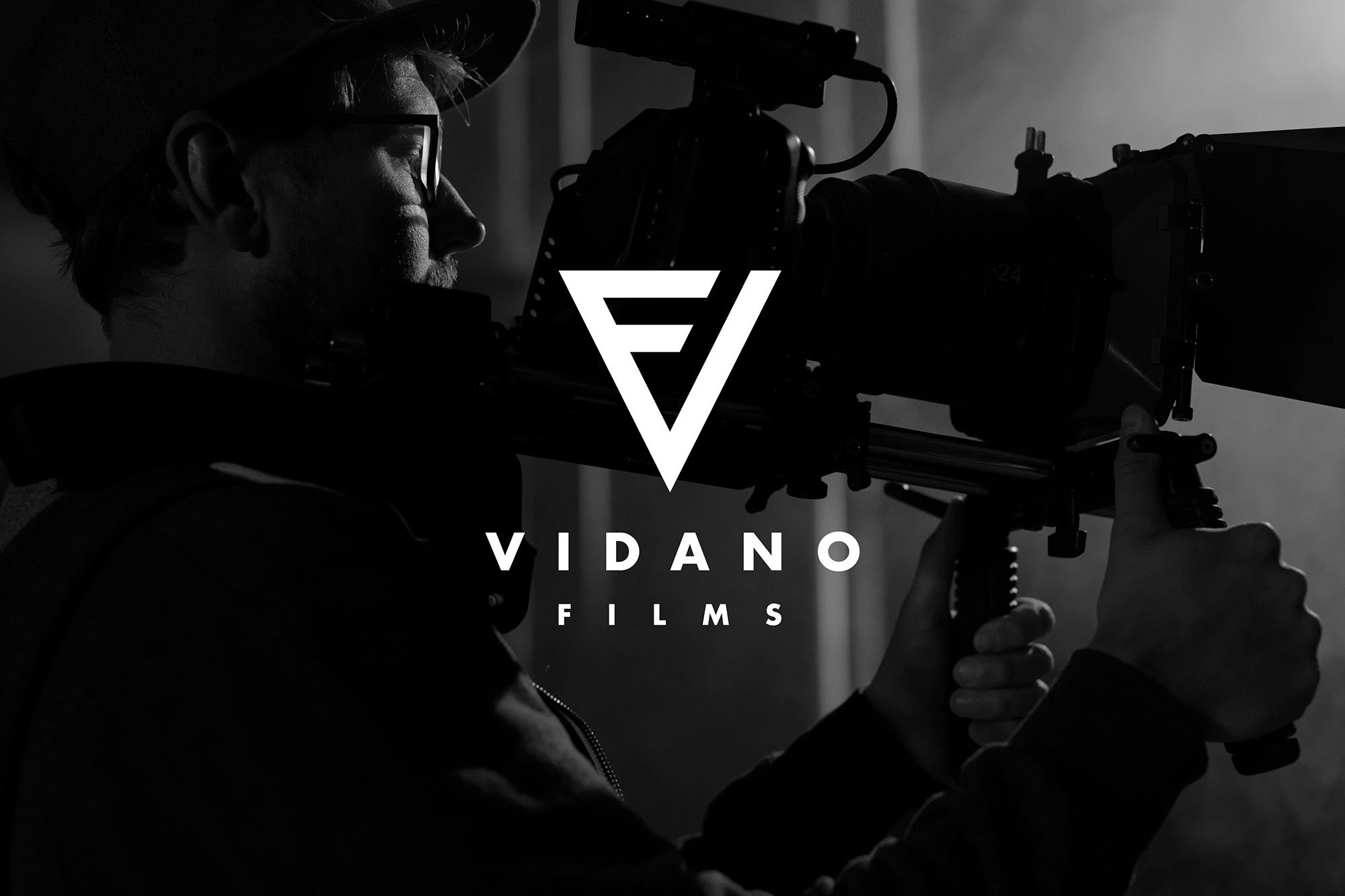 A black and white image of a person wearing glasses and a hat, looking through a camera or telescope, with the logo of Vidano Films overlayed in the center.
